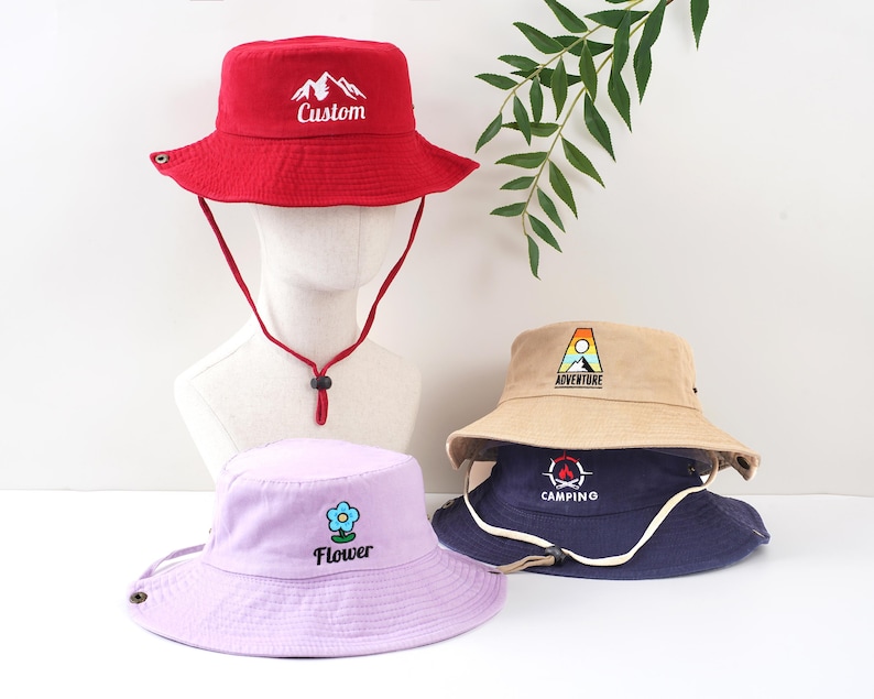K&ouml;nnte beinhalten: Eine Auswahl an Fischerh&uuml;ten in verschiedenen Farben, darunter Rot, Beige, Marineblau und Lavendel. Jeder Hut hat ein gesticktes Design, wie eine Berglandschaft, eine Blume oder die W&ouml;rter "Adventure" oder "Camping". Die H&uuml;te haben Kinnriemen.