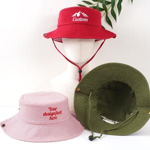 K&ouml;nnte beinhalten: Drei Fischerh&uuml;te in Rot, Rosa und Olivgr&uuml;n. Der rote Hut hat den gestickten Schriftzug "Custom" mit einem Bergmotiv auf der Vorderseite. Der rosa Hut hat den gestickten Schriftzug "Your design/text here". Alle H&uuml;te haben einen Kinnriemen.