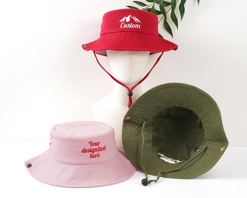 K&ouml;nnte beinhalten: Drei Fischerh&uuml;te in Rot, Rosa und Olivgr&uuml;n. Der rote Hut hat den gestickten Schriftzug "Custom" mit einem Bergmotiv auf der Vorderseite. Der rosa Hut hat den gestickten Schriftzug "Your design/text here". Alle H&uuml;te haben einen Kinnriemen.
