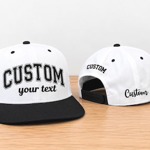 May include: Two white snapback caps with black brims. The front cap features the word CUSTOM in a bold block font with the text your text below it. The rear cap displays the word Custom in a script font on the side and back panels.