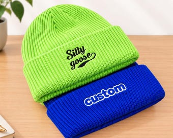 May include: A bright lime green ribbed knit beanie featuring the black embroidered text Silly goose and a royal blue ribbed knit beanie featuring the white embroidered text custom stacked underneath the green beanie.