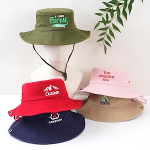 K&ouml;nnte beinhalten: Eine Auswahl an Fischerh&uuml;ten in verschiedenen Farben, darunter Gr&uuml;n, Rot, Rosa und Marineblau. Jeder Hut hat gestickten Text oder eine Grafik, wie "I Love Forest Hiking", eine Berglandschaft oder das Wort "Camping".
