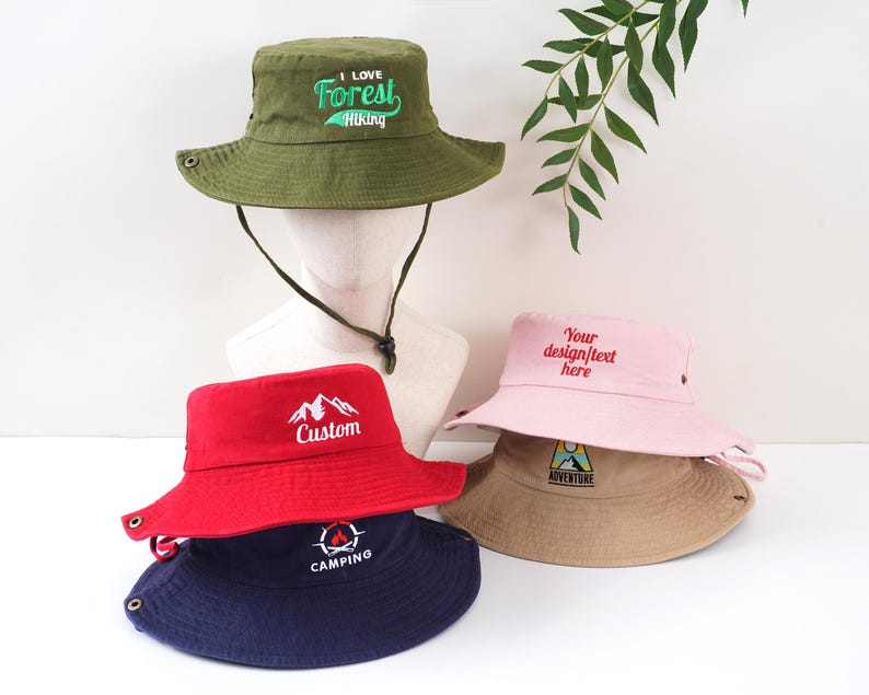 K&ouml;nnte beinhalten: Eine Auswahl an Fischerh&uuml;ten in verschiedenen Farben, darunter Gr&uuml;n, Rot, Rosa und Marineblau. Jeder Hut hat gestickten Text oder eine Grafik, wie "I Love Forest Hiking", eine Berglandschaft oder das Wort "Camping".
