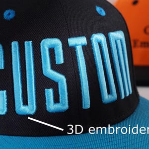 May include: Black and blue baseball cap with "CUSTOM" embroidered in light blue 3D lettering. The cap features a flat brim and a matching blue top button. The text "3D embroidery" is also visible.