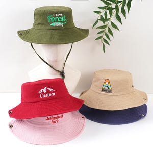 K&ouml;nnte beinhalten: Eine Sammlung von Fischerh&uuml;ten in verschiedenen Farben, darunter Olivgr&uuml;n, Rot, Beige und Rosa. Der olivgr&uuml;ne Hut hat die Aufschrift "I Love Forest Hiking". Der rote Hut hat eine Berg-Grafik und das Wort "Custom". Der beige Hut hat ein "Adventure"-Logo.