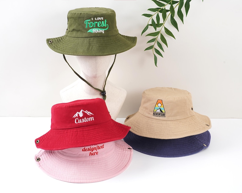 K&ouml;nnte beinhalten: Eine Sammlung von Fischerh&uuml;ten in verschiedenen Farben, darunter Olivgr&uuml;n, Rot, Beige und Rosa. Der olivgr&uuml;ne Hut hat die Aufschrift "I Love Forest Hiking". Der rote Hut hat eine Berg-Grafik und das Wort "Custom". Der beige Hut hat ein "Adventure"-Logo.