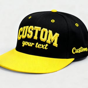 May include: Black and yellow baseball cap with the word "CUSTOM" embroidered in yellow across the front. The text "your text" is embroidered below "CUSTOM". The brim is yellow, and the word "Custom" is embroidered on the side.