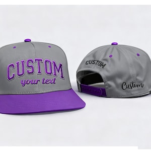 May include: A gray baseball cap with a purple brim and a purple button on top. The word "CUSTOM" is embroidered in purple on the front, with "your text" below it. The back of the cap also has the word "CUSTOM" embroidered.