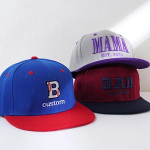 May include: Three baseball caps, one blue with a red brim and a white letter "B" with a baseball stitch design, one burgundy with a navy brim and the word "DAD" in purple with the text "EST. 2020", and one gray with a purple brim and the word "MAMA" in purple with the text "EST. 2025".