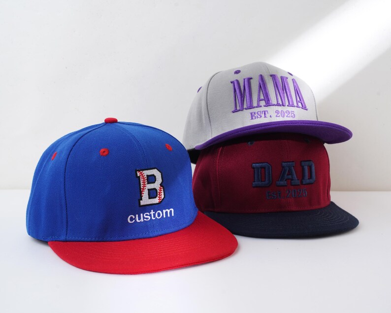 May include: Three baseball caps, one blue with a red brim and a white letter "B" with a baseball stitch design, one burgundy with a navy brim and the word "DAD" in purple with the text "EST. 2020", and one gray with a purple brim and the word "MAMA" in purple with the text "EST. 2025".