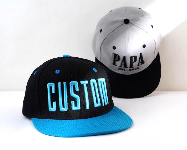 May include: Two baseball caps. One black cap with "CUSTOM" in blue lettering and a blue brim. The other is a gray cap with "PAPA EST. 2025" in black lettering and a black brim. Both caps have a flat bill.