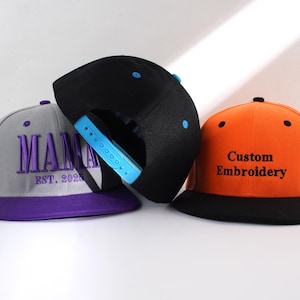 May include: Three baseball caps, one gray with purple trim and the text "MAMA EST. 2025" embroidered in purple, one black with a blue snap, and one orange with a black brim and the text "Custom Embroidery" embroidered in black.