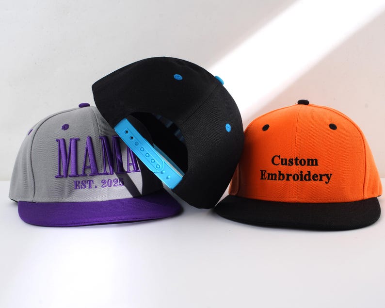 May include: Three baseball caps, one gray with purple trim and the text "MAMA EST. 2025" embroidered in purple, one black with a blue snap, and one orange with a black brim and the text "Custom Embroidery" embroidered in black.