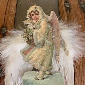 Könnte beinhalten: Vintage Engel-Weihnachtsschmuck mit goldenem Aufhängeband. Der Engel trägt einen langen Mantel und hält einen Korb mit Blumen, mit weißen Federflügeln. Die Flügel sind an einem Holzgrund befestigt.