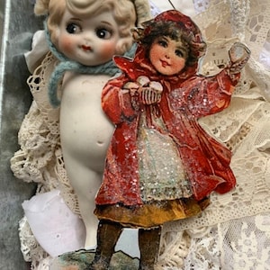 Puede incluir: Una muñeca de porcelana vintage con un gorro azul y un adorno de papel de una niña con un abrigo rojo que sostiene una bola de nieve. El adorno tiene un acabado brillante y está colgado de un gancho plateado.