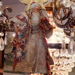 Pode incluir: Um enfeite de Natal em estilo vintage com o Pai Natal, adornado com glitter e uma estrela dourada. O enfeite tem uma longa barba branca, um chapéu vermelho e um casaco detalhado. Está pendurado numa candelabro com detalhes em cristal.