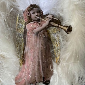 May include: A vintage-style angel ornament with gold glitter wings, playing a trumpet. The angel is wearing a pink dress and has a white halo.
