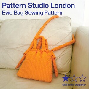May include: An orange Evie bag sewing pattern from Pattern Studio London. The bag is made of a textured fabric and has a drawstring closure. The bag has long straps and is displayed on a cream-colored sofa. The skill level is beginner.