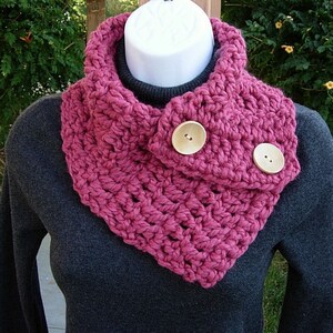 May include: A pink crocheted cowl with two large wooden buttons. The cowl is worn on a gray sweater.
