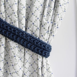 May include: A blue crocheted curtain tieback, tied around a white curtain with a floral pattern.