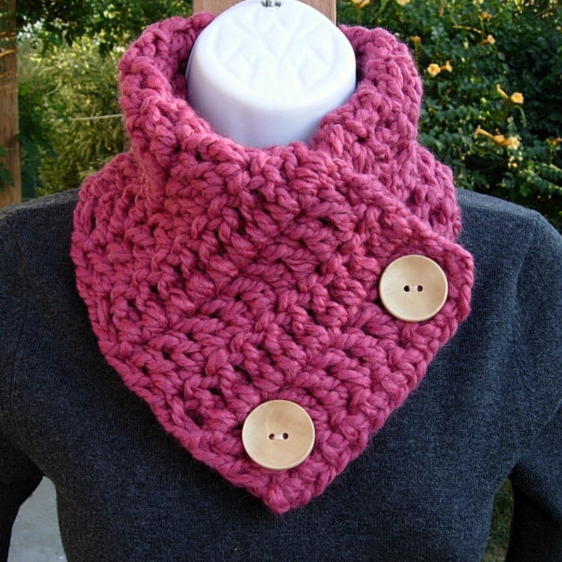 May include: A pink crocheted cowl with two wooden buttons. The cowl is folded over and the buttons are on the fold.