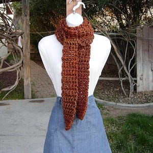 May include: A brown crocheted scarf with a lacy pattern. The scarf is displayed on a mannequin wearing a white shirt and a denim skirt.