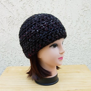May include: A black crocheted beanie with flecks of pink and white yarn. The beanie is on a mannequin head with brown hair.