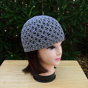 May include: A grey crocheted beanie hat with a simple, open stitch pattern. The hat is shown on a mannequin head with brown hair.