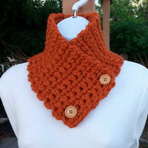 May include: A crocheted orange neck warmer with two wooden buttons. The neck warmer is made with a textured stitch and has a triangular shape.