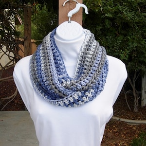 May include: A crocheted infinity scarf in shades of blue, grey, and white. The scarf is made with a chunky yarn and has a simple, textured stitch.