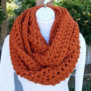 May include: A chunky knit infinity scarf in a warm burnt orange color. The scarf is made with a thick yarn and has a simple, textured stitch pattern.