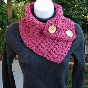 May include: A pink crocheted cowl with two wooden buttons. The cowl is worn over a gray sweater.