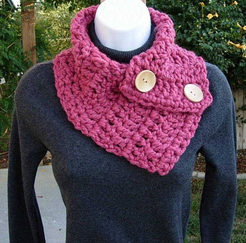May include: A pink crocheted cowl with two wooden buttons. The cowl is worn over a gray sweater.