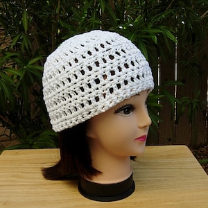 May include: White crocheted beanie hat with a lacy open stitch pattern. The hat is on a mannequin head with short brown hair.