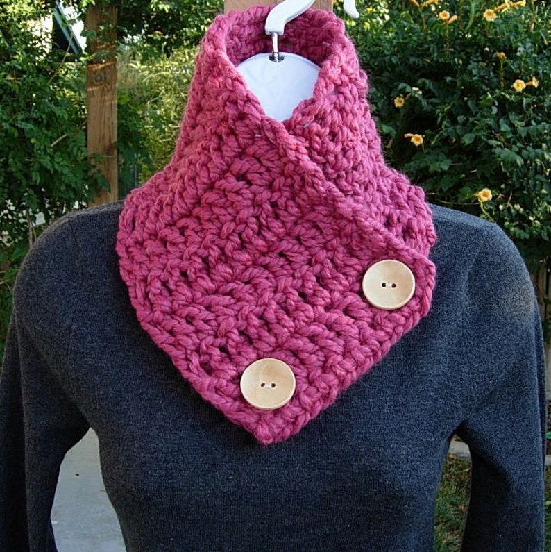 May include: A pink crocheted cowl with two wooden buttons. The cowl is folded over and worn around the neck.
