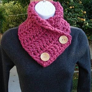 May include: A pink crocheted cowl with two wooden buttons. The cowl is worn over a gray sweater.