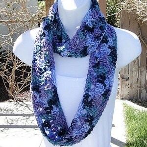 May include: A handmade infinity scarf in shades of blue, purple, and teal. The crochet scarf is displayed on a white mannequin. The yarn has a textured appearance, with a mix of light and dark tones.