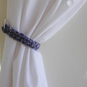 May include: A crocheted curtain tieback in shades of purple and gray, holding white curtains. The handmade tieback is a simple, yet elegant home decor item. The soft texture of the yarn contrasts with the smooth fabric of the curtains.