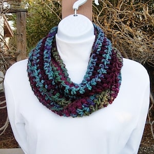 May include: A crocheted infinity scarf in shades of purple, teal, and green. The scarf is made with a chunky yarn and has a simple, textured stitch.