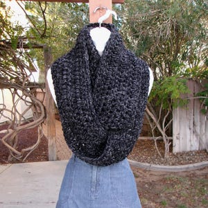 以下が含まれることがあります： 黒とグレーの色合いの手編みの分厚いインフィニティスカーフ。白いマネキンの頭に展示され、デニムスカートの上に掛けられています。太い糸が、肌触りの良い、居心地の良い外観を作り出し、寒い季節に最適です。周囲約150cm。