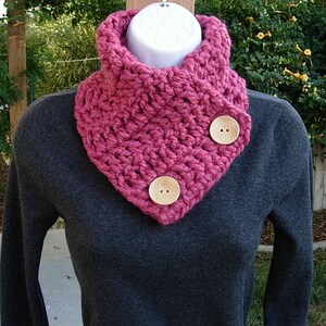May include: A pink crocheted cowl with two wooden buttons. The cowl is folded over and worn around the neck.