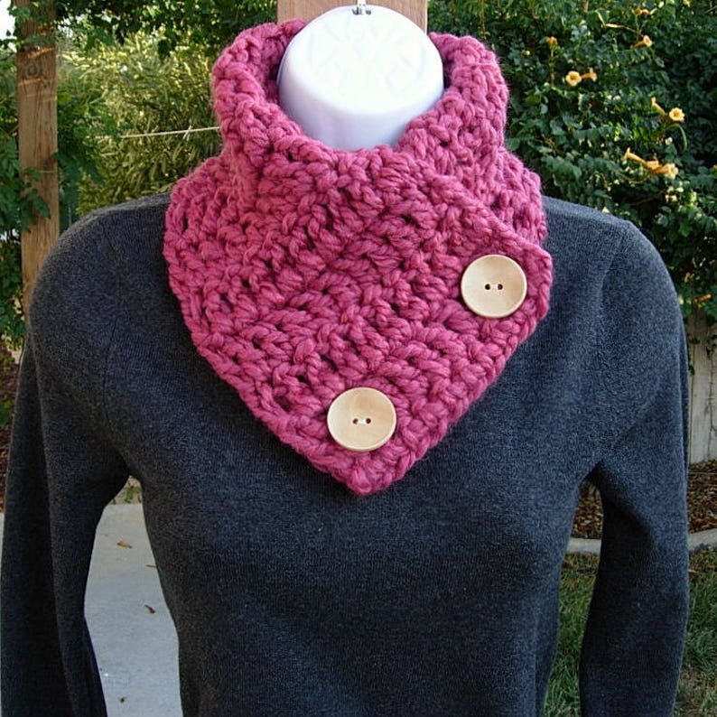 May include: A pink crocheted cowl with two wooden buttons. The cowl is folded over and worn around the neck.