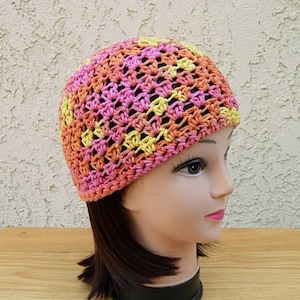 May include: A handmade crochet beanie in a mix of pink, yellow, and orange. The hat is made with a loose, open weave, and is shown on a mannequin head. The beanie is a colorful accessory.