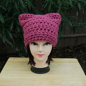 May include: A pink crocheted hat with cat ears. The hat is on a mannequin head.