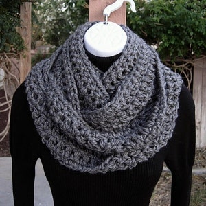May include: A chunky, dark gray infinity scarf featuring a thick, textured crochet knit pattern. The scarf is draped in multiple loops around a black long-sleeve shirt, showcasing a dense, interconnected yarn construction with visible stitch details.