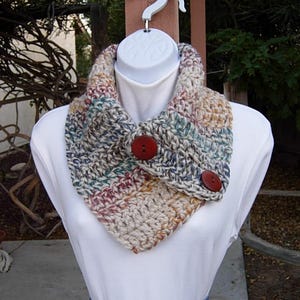 May include: A crocheted cowl with a multi-colored pattern in shades of brown, beige, red, and blue. The cowl has two large brown buttons.