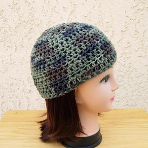 May include: A crocheted beanie hat in shades of green, blue, and brown. The hat is made with a loose, open weave, and is shown on a mannequin head with brown hair.