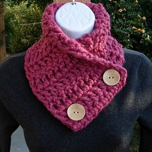 May include: A pink crocheted cowl with two wooden buttons. The cowl is folded over and the buttons are on the side.