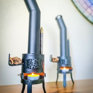 Puede incluir: Dos pequeñas estufas de velas de metal negro. Cada una tiene un cuerpo cilíndrico, una chimenea en ángulo y tres patas. Una tiene una pequeña bandeja con trozos de madera. Una vela encendida está dentro de cada estufa.