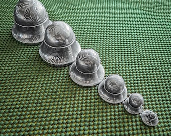 May include: A collection of six silver-colored, hat-shaped objects with a textured, swirling pattern. The objects are arranged in a diagonal line, decreasing in size from left to right. The background is a textured green surface.